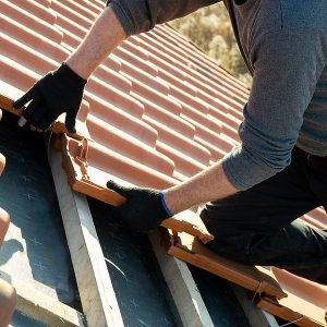 Closeup of worker hands installing yellow ceramic roofing tiles mounted on wooden boards covering residential building roof under construction.