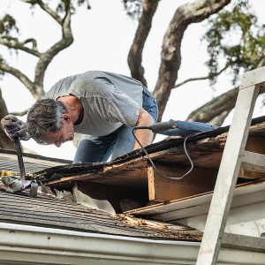 Man using crowbar to remove rotten wood from leaky roof. After removing fascia boards he has discovered that the leak has extended into the beams and decking.